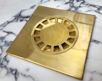 May include: A square, gold-colored drain cover with a circular, decorative grate. The drain cover is made of brass and has a polished finish.