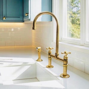 Peut inclure: Un robinet de cuisine doré avec un bec en arc de cercle et des poignées en forme de croix. Le robinet est monté sur un plan de travail blanc, avec un pulvérisateur latéral assorti. Le fond comprend des carreaux de métro blancs et des armoires bleu sarcelle.