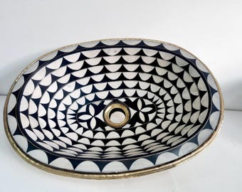 Op de afbeelding: Ovale keramische wastafel met een zwart-wit geometrisch patroon. De wastafel heeft een herhalend ontwerp van zwarte driehoeken en witte halve cirkels, met een goudkleurige rand en afvoer. Het centrale ontwerp is bloemvormig.