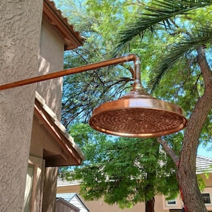 May include: A copper outdoor shower head with a hammered finish, mounted on a copper pipe against a stucco wall. The shower head is positioned under a tree with green leaves.
