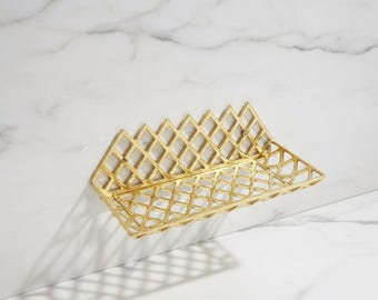 May include: A gold-colored, wall-mounted basket with a geometric diamond pattern. The basket has a rectangular shape with a sloped top edge. The basket is made of metal and is mounted on a white marble wall.