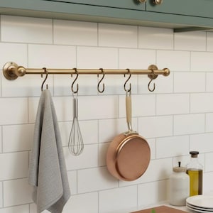 May include: A brass kitchen rail with six hooks, displaying a grey towel, a whisk, and a copper-bottomed pan. The rail is mounted on a white tiled wall, with a green cabinet above.