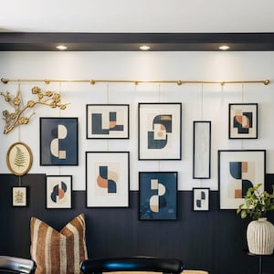 May include: A gallery wall arrangement featuring multiple framed prints with abstract geometric shapes in navy, cream, and terracotta. A gold metal rod hangs the frames, accompanied by a gold branch sculpture and a fern print in an oval frame.