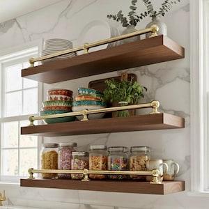 Könnte beinhalten: Dreistufige Holzregale mit Messinggeländern an einer Wand mit Marmormuster. Die Regale zeigen Tellerstapel, dekorative Schalen, Gläser mit Lebensmitteln und Tassen. Die Regale sind dunkelbraun.