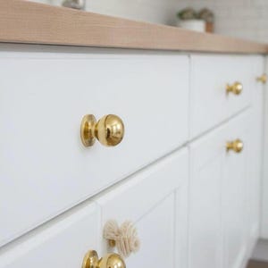 K&ouml;nnte beinhalten: Nahaufnahme von wei&szlig;en K&uuml;chenschr&auml;nken mit goldfarbenen, kugelf&ouml;rmigen Kn&ouml;pfen. Die Schr&auml;nke sind sauber und modern, mit einer hellen Holz-Arbeitsplatte im Hintergrund. Die Kn&ouml;pfe verleihen dem K&uuml;chendesign einen Hauch von Eleganz.