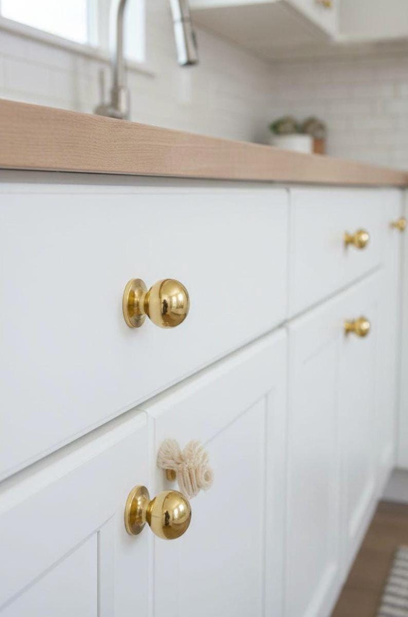 K&ouml;nnte beinhalten: Nahaufnahme von wei&szlig;en K&uuml;chenschr&auml;nken mit goldfarbenen, kugelf&ouml;rmigen Kn&ouml;pfen. Die Schr&auml;nke sind sauber und modern, mit einer hellen Holz-Arbeitsplatte im Hintergrund. Die Kn&ouml;pfe verleihen dem K&uuml;chendesign einen Hauch von Eleganz.