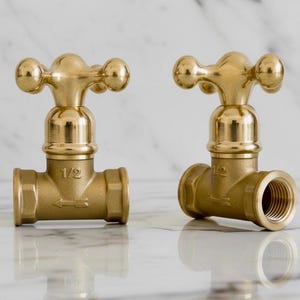 May include: Two brass-colored faucet handles with a classic design. Each handle has a cross-shaped top with rounded ends and a cylindrical base. The handles are labeled "1/2" and are set against a white marble background.