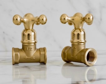 Peut inclure: Deux robinets en laiton de conception classique. Chaque robinet a un dessus en forme de croix avec des extrémités arrondies et une base cylindrique. Les robinets sont marqués "1/2" et ont une flèche indiquant le sens d'écoulement. Sur fond de marbre.