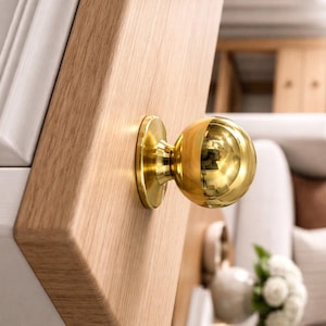 Peut inclure: Gros plan d'un bouton de meuble sphérique doré fixé sur une surface en bois clair. Le bouton a une base ronde et une finition polie et réfléchissante. Le grain du bois est visible sur la surface.
