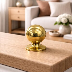 May include: A polished gold sphere ornament with a circular base, reflecting the room's details. It sits on a light-coloured wooden surface. This decorative item could be used as a door knob or furniture accent, adding a touch of elegance.