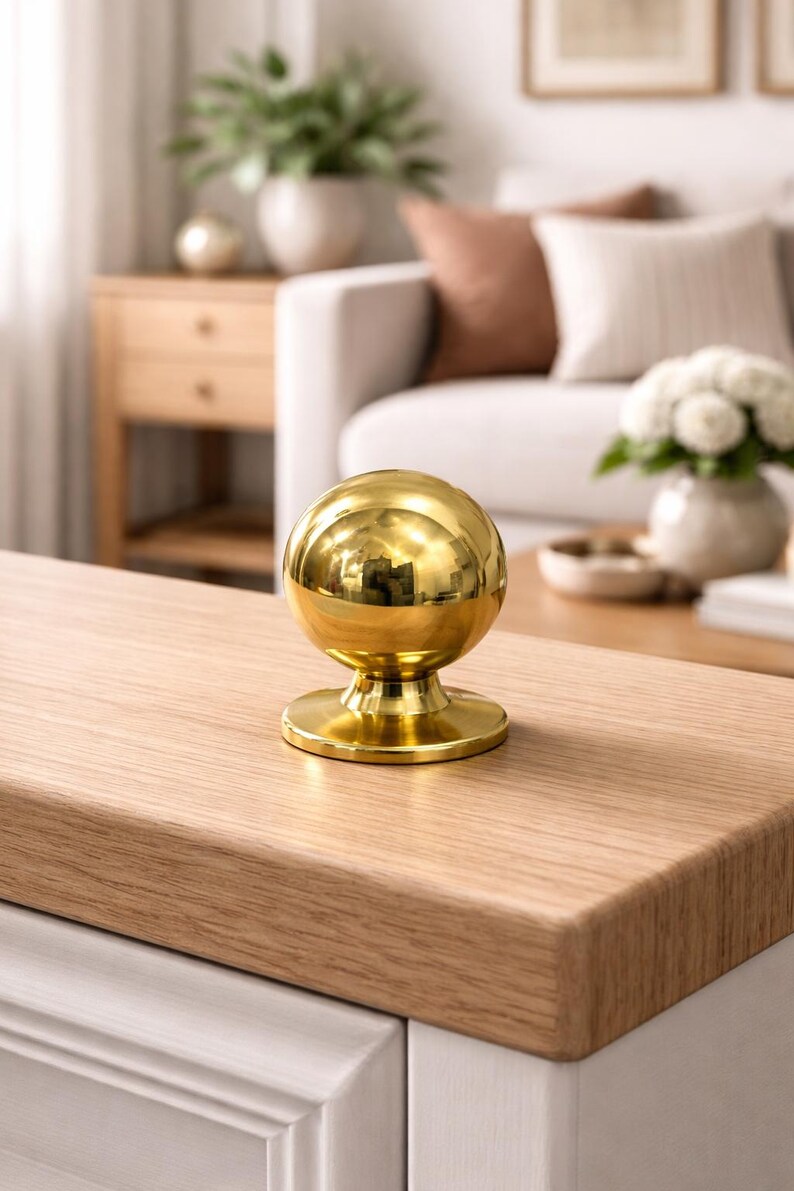 May include: A polished gold sphere ornament with a circular base, reflecting the room's details. It sits on a light-coloured wooden surface. This decorative item could be used as a door knob or furniture accent, adding a touch of elegance.