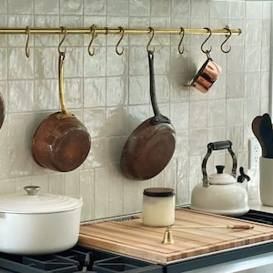 Peut inclure: Une cuisine avec un dosseret en carrelage blanc et une barre en laiton avec des crochets sur lesquels pendent des casseroles et des poêles en cuivre. Une casserole en céramique blanche repose sur une planche à découper en bois sur une cuisinière noire.