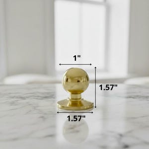 K&ouml;nnte beinhalten: Ein goldfarbener Schrankknauf mit kugelf&ouml;rmiger Oberseite und kreisf&ouml;rmiger Basis. Der Knauf ist 2,54 cm breit und 4 cm hoch. Die Hardware wird auf einer wei&szlig;en Marmoroberfl&auml;che gezeigt.