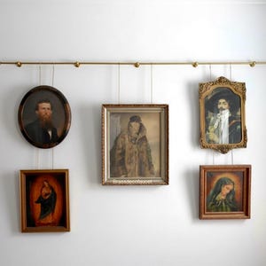 Könnte beinhalten: Eine Sammlung gerahmter Vintage-Porträts und religiöser Kunst, die an einer Messingstange an einer weißen Wand hängen. Die Rahmen variieren in Form und Stil, mit Porträts und religiösen Figuren.