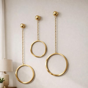 Può includere: Tre elementi di arte murale in metallo color oro. Ogni pezzo presenta un anello circolare sospeso da una catena a un supporto a parete rotondo color oro. Gli anelli variano di dimensioni, creando un effetto a strati. Il muro è di colore neutro.