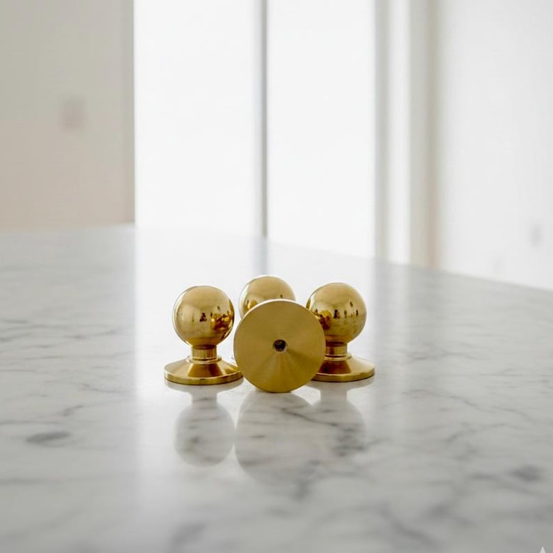 K&ouml;nnte beinhalten: Vier Messing-T&uuml;rkn&ouml;pfe mit kugelf&ouml;rmigem Design und kreisf&ouml;rmiger Basis, angeordnet auf einer Marmoroberfl&auml;che. Die Kn&ouml;pfe haben eine gl&auml;nzende, reflektierende Oberfl&auml;che. Der Hintergrund ist unscharf.