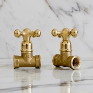 Peut inclure: Deux robinets en laiton avec une finition polie. Chaque robinet a une poignée en forme de croix et une sortie filetée. Les robinets sont marqués "1/2" et sont posés sur un fond en marbre.