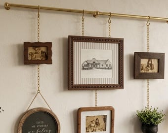 May include: A gold-toned metal rod mounted horizontally with four gold chains hanging from S-hooks. The chains support three rectangular wooden frames, one circular wooden frame with the text "THIS ME STORIES we belong", and a small black planter.