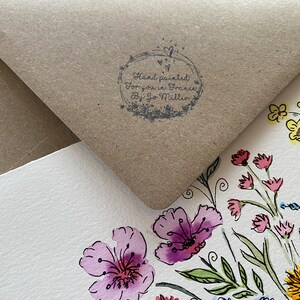 May include: A white card with a hand-painted floral design in watercolor. The card features a variety of flowers in pink, yellow, blue, and green. The card is partially covered by a brown envelope with a stamp that reads "Hand painted for you in France by Jo Mitton".
