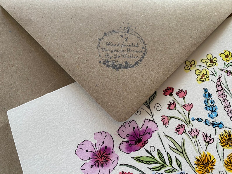 May include: A white card with a hand-painted floral design in watercolor. The card features a variety of flowers in pink, yellow, blue, and green. The card is partially covered by a brown envelope with a stamp that reads "Hand painted for you in France by Jo Mitton".