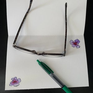 May include: A white card with a green pen, a pair of tortoiseshell glasses, and two purple flowers drawn on it.