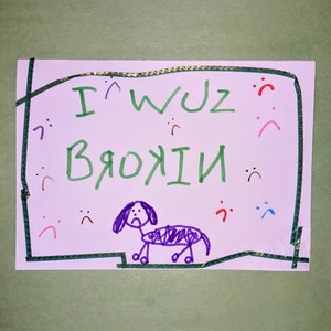 May include: A purple drawing of a dog with a sad face. The dog is on a white background with green and gold borders. The text "I wuz broke" is written in green above the dog.