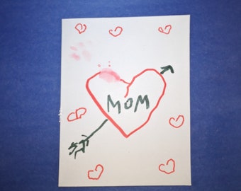 May include: A white card with a red heart drawn on it. The heart has an arrow through it and the word "MOM" written inside. There are several red hearts drawn around the large heart.