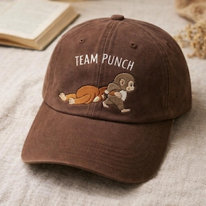 May include: A brown baseball cap with the embroidered text "TEAM PUNCH" above a cartoon monkey design. The design features two monkeys, one lying down and the other standing. The cap has a curved brim and a button on top.