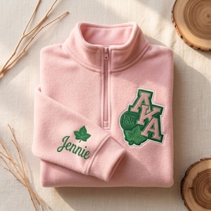 May include: A light pink fleece pullover with a quarter-zip closure. The left sleeve cuff has the name "Jennie" embroidered in green with a green leaf. The chest features a green and pink embroidered patch with the letters "AKA" and "EST 1908".