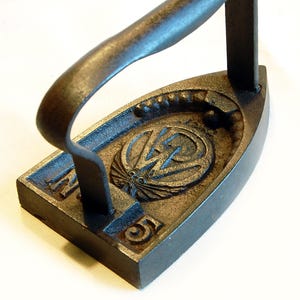 Könnte beinhalten: Antikes Bügeleisen aus Gusseisen mit gebogenem Griff. Das Bügeleisen ist dunkelbraun mit der Nummer "5" und einem dekorativen Emblem auf der Oberseite. Das Emblem scheint ein stilisiertes Design mit einem Blitz zu sein.