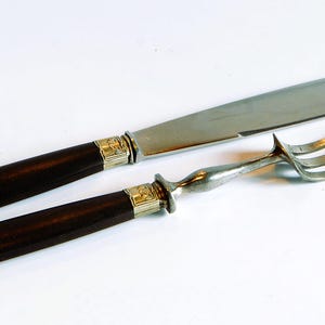 Könnte beinhalten: Ein Messer- und Gabel-Set mit dunkelbraunen Griffen und silberfarbenen Klingen und Zinken. Die Griffe haben goldfarbene Akzente an den Enden. Die Messerklinge ist lang und schlank. Die Gabel hat drei Zinken.