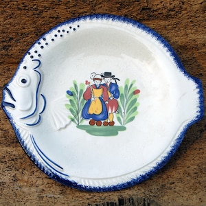 Peut inclure: Une assiette en céramique blanche en forme de poisson avec une bordure bleue et blanche. L'assiette présente une scène peinte de deux personnes en costumes traditionnels debout devant un buisson vert.