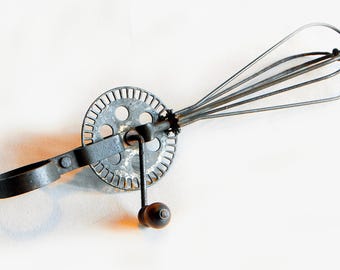 Può includere: Un frullino vintage a manovella con una frusta in metallo e un ingranaggio rotondo. Il frullino è in metallo e ha una finitura argentata.