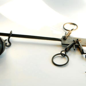 Könnte beinhalten: Antike schwarze Metallwaage mit rundem Gewicht und gebogenem Haken. Die Waage hat einen langen Arm und mehrere Ringe zum Aufhängen von Gegenständen. Sie dient wahrscheinlich zum Messen von Gewicht.