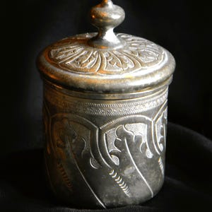 Puede incluir: Un tarro antiguo de color plateado con tapa. El tarro y la tapa están adornados con intrincados diseños florales y de hojas. La tapa tiene un pequeño pomo redondeado. El tarro es cilíndrico y se asienta sobre un fondo oscuro.