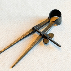 Könnte beinhalten: Ein antiker Kompass aus Metall mit schwarzer Oberfläche und einem Schmetterlings-Einstellknopf. Der Kompass hat zwei spitze Beine und einen runden Kopf.