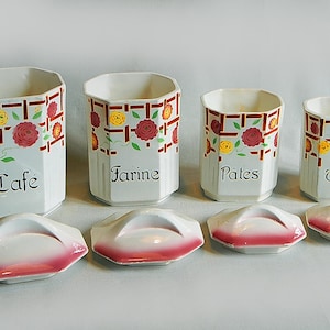 May include: Set of four white ceramic canisters with lids. Each canister has a floral design with red, yellow, and green accents. The canisters are labeled "Café", "Farine", "Pâtes", and "Thé".