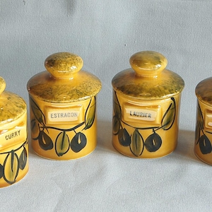 Pode incluir: Quatro potes de especiarias de cerâmica amarela com tampas. Cada pote tem um rótulo com o nome de uma especiaria: Curry, Estragão, Louro e Tomilho. Os potes são decorados com desenhos de ramos de oliveira.