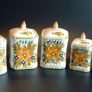 Könnte beinhalten: Ein Set aus fünf weißen Keramikdosen mit Deckeln, verziert mit handgemalten orangefarbenen Blumen, grünen Blättern und blauen Akzenten. Die Dosen sind auf der Vorderseite mit den Wörtern CAFE, FARINE, SUCRE, POIVRE und SEL in schwarzer Schrift beschriftet.