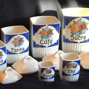 Peut inclure: Ensemble de pots de cuisine en céramique blanche avec des motifs bleus et floraux. Chaque pot est muni d'un couvercle et porte des inscriptions françaises comme "Café" et "Sucre". L'ensemble est présenté sur fond sombre.