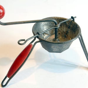 Może przedstawiać: Vintage metalowy przecierak do warzyw z szarą, stożkową misą z wytłoczonym tekstem, obrotową korbką z czerwoną drewnianą gałką oraz bocznym uchwytem z czerwoną drewnianą rączką. Urządzenie stoi na trzech metalowych nóżkach z małymi stopkami.