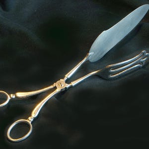 Puede incluir: Servidor de pastel plateado con una hoja de cuchillo y un extremo de tenedor, conectado por un mecanismo de tijera. El servidor tiene dos lazos para los dedos y está sobre un fondo oscuro. Un utensilio de servicio clásico.