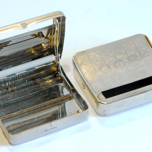 May include: Two vintage silver metal cigarette cases. One case is open, revealing the interior with a white roller and metal dividers. The other case is closed, with a geometric pattern on the lid. Both cases have a rectangular shape.
