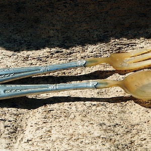 Può includere: Un set vintage di forchetta e cucchiaio. I manici sono color argento con un motivo floreale e le superfici per mangiare sono di colore ambra traslucido. Il set è esposto su una superficie strutturata marrone chiaro.