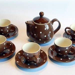 Op de afbeelding: Een bruine keramische theeset met witte stippen. De set bevat een theepot met deksel en zes theekopjes met schotels. De kopjes en de theepot hebben een glanzende afwerking. De binnenkant van de kopjes is crèmekleurig.