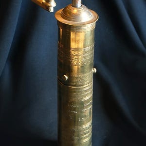 May include: A vintage brass pepper mill with a cylindrical body and a crank handle. The mill features decorative etched bands and a small handle on the side. The brass has a warm, aged patina, suggesting its use over time. The mill is designed for grinding peppercorns.