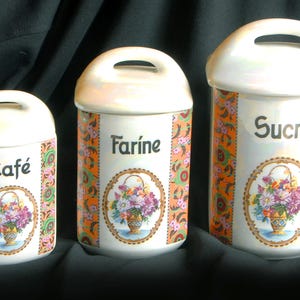 Puede incluir: Juego de tres botes de cerámica con tapas y asas blancas. Cada bote tiene un diseño floral y una etiqueta: "Café", "Farine" y "Sucre". Los botes tienen un colorido estampado naranja y floral en los laterales.