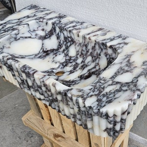 May include: A white marble sink with black veining. The sink has a unique, wavy design and a rippled edge. The sink is sitting on a wooden pallet.