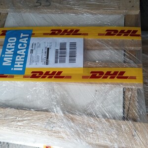 May include: A wooden pallet wrapped in clear plastic with yellow tape that says "DHL" in red letters. A blue label with the text "MİKRO İHRACAT" is attached to the pallet.