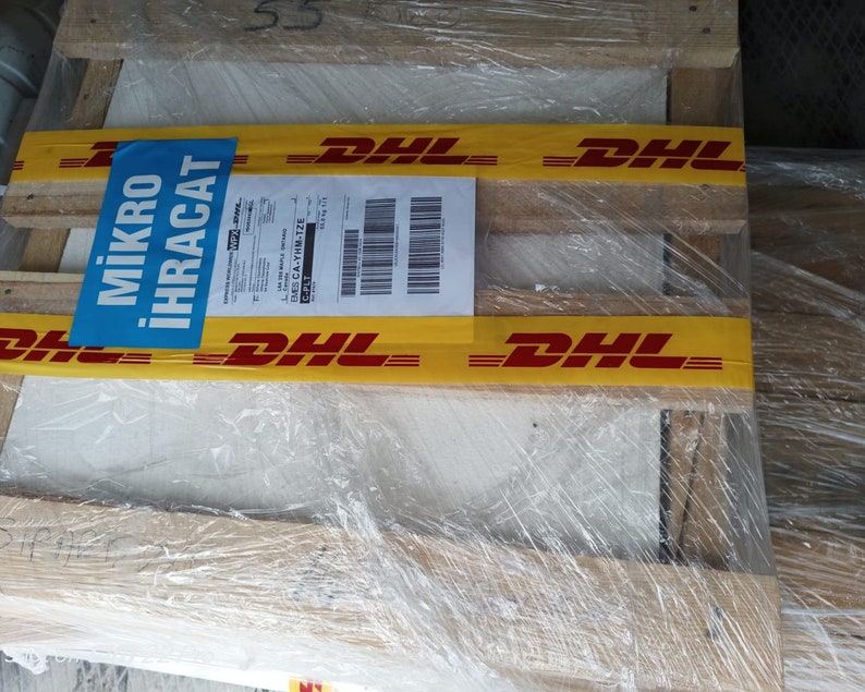 May include: A wooden pallet wrapped in clear plastic with yellow tape that says "DHL" in red letters. A blue label with the text "MİKRO İHRACAT" is attached to the pallet.